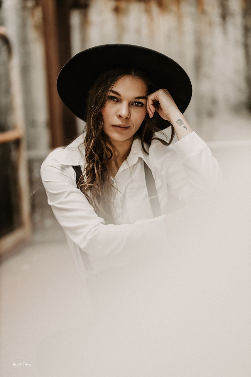 A portrait of someone wearing a wide-brimmed black hat and white button-down shirt in soft, moody lighting.
