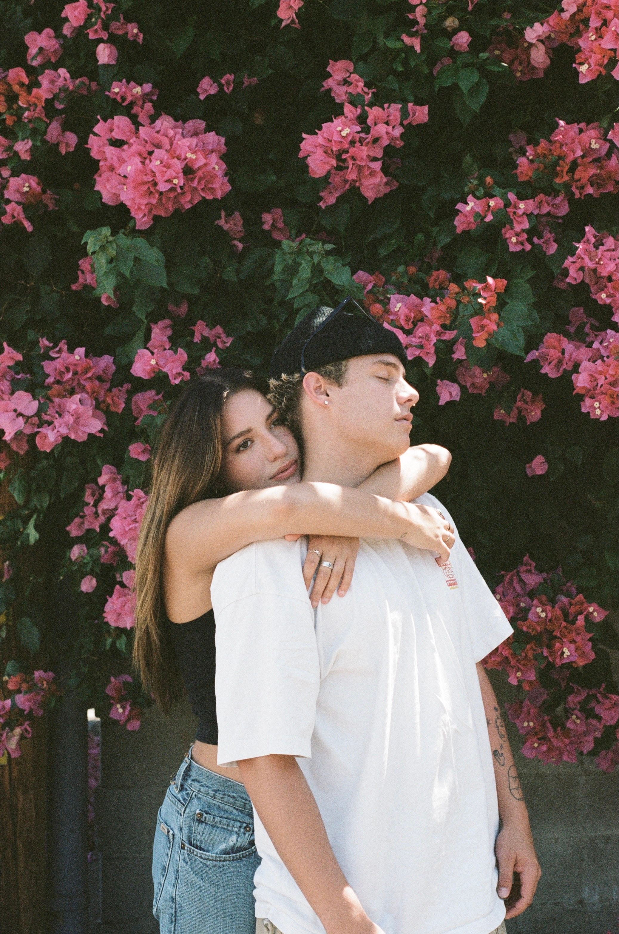 A romantic couple embracing in front of pink bougainvillea flowers on a dark background.