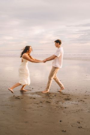 A couple dancing playfully together on a beach at sunset wearing light colored outfits.