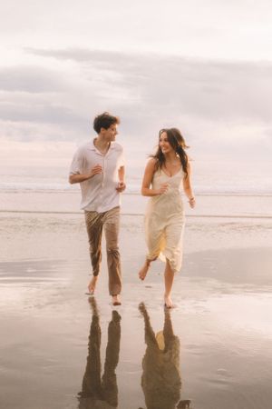 A couple running joyfully along the wet sand at sunset with their reflections visible in the water.