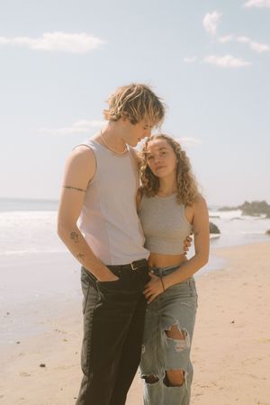 A romantic couple embracing on a sunny beach, wearing casual outfits with a white tank top and ripped jeans.