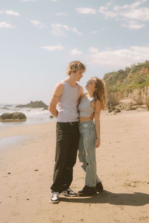 A couple in casual attire sharing an intimate moment on a sandy beach with rocky cliffs in the background.
