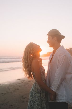 Romantic couple laughing together on beach at sunset in soft pink light.