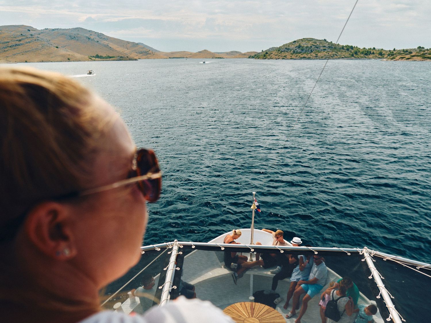 Someone wearing sunglasses looks out over blue waters from the bow of a boat near mountainous coastline on a sunny day.