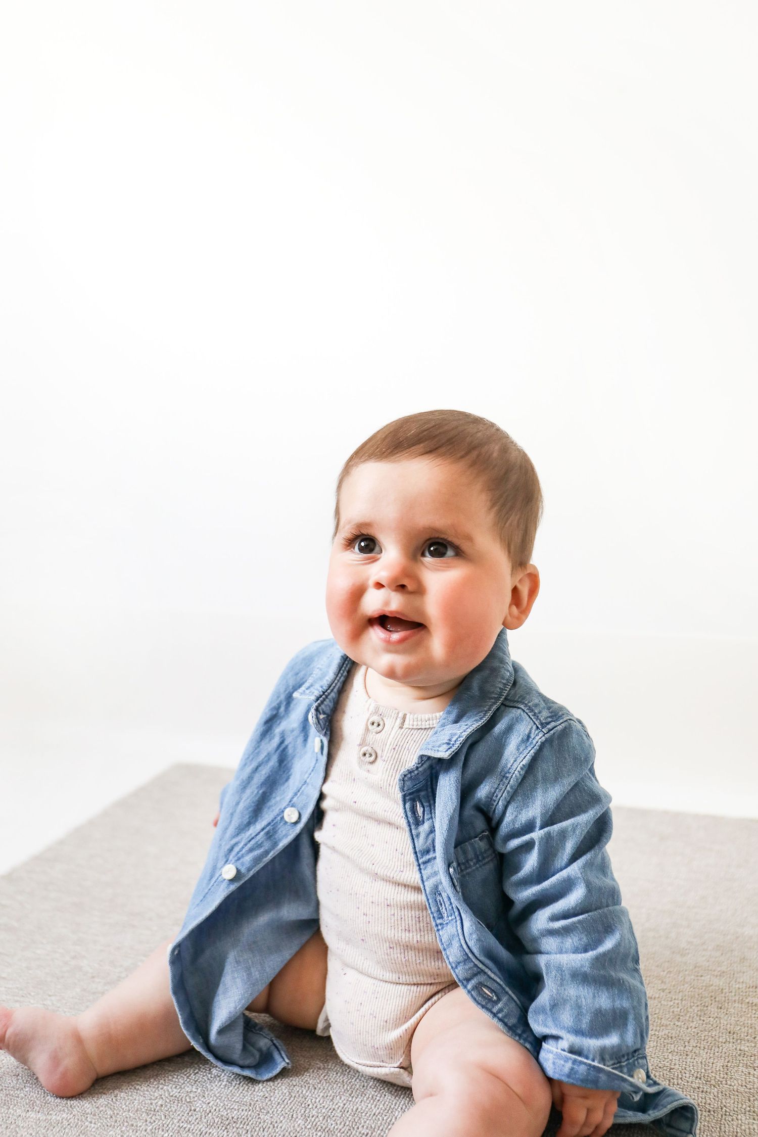 Baby wearing white onesie and denim jacket sits on grey surface against white background, during Sitter photoshoot based in Nottingham