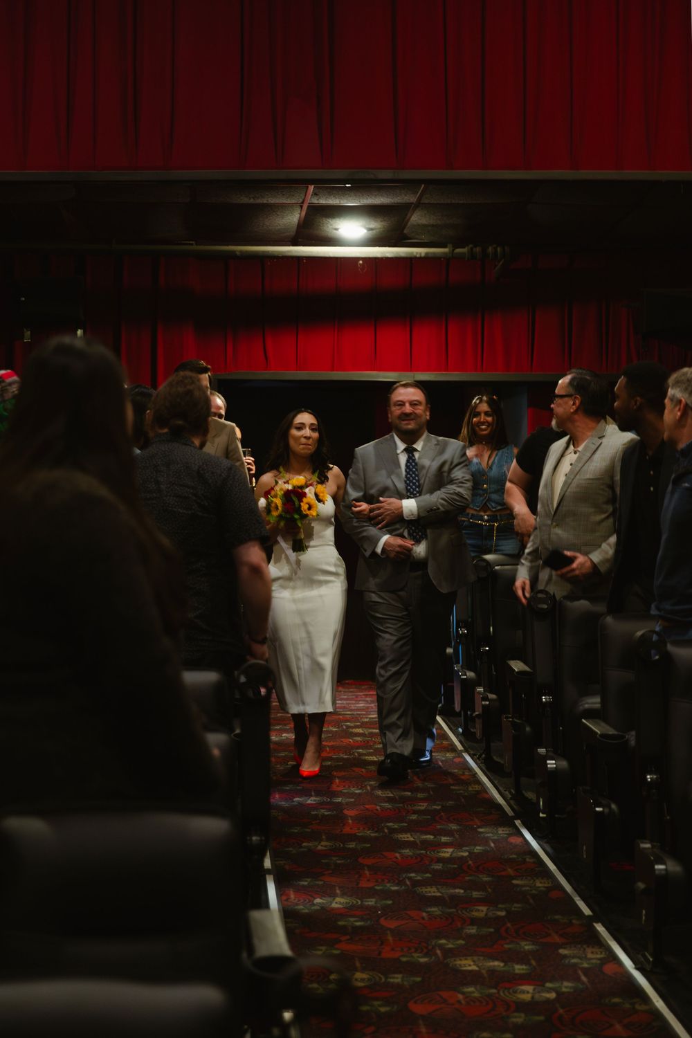 A wedding ceremony procession walking down the aisle of a theater with red curtains.