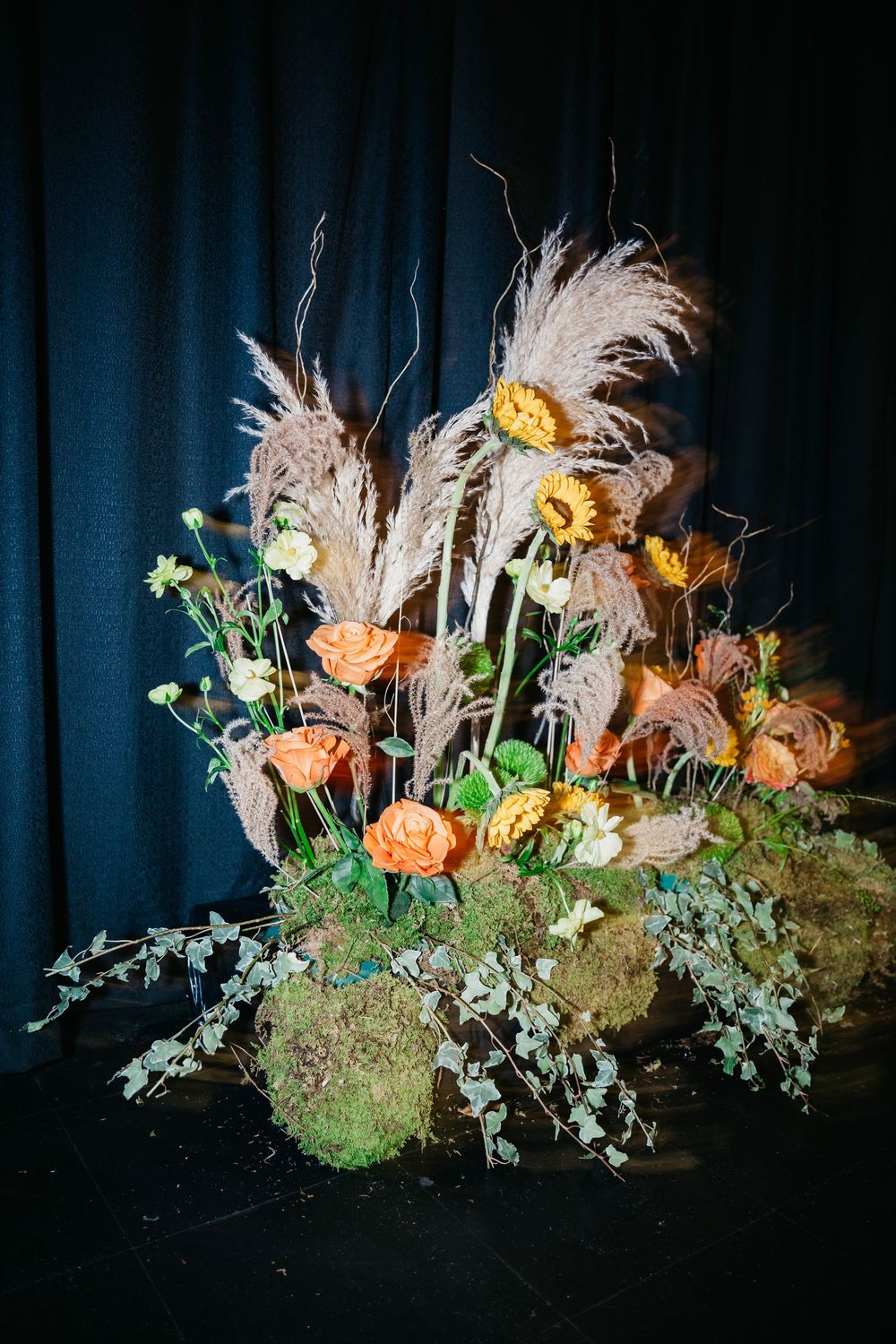A dramatic floral arrangement with orange blooms and pampas grass against a dark blue backdrop.
