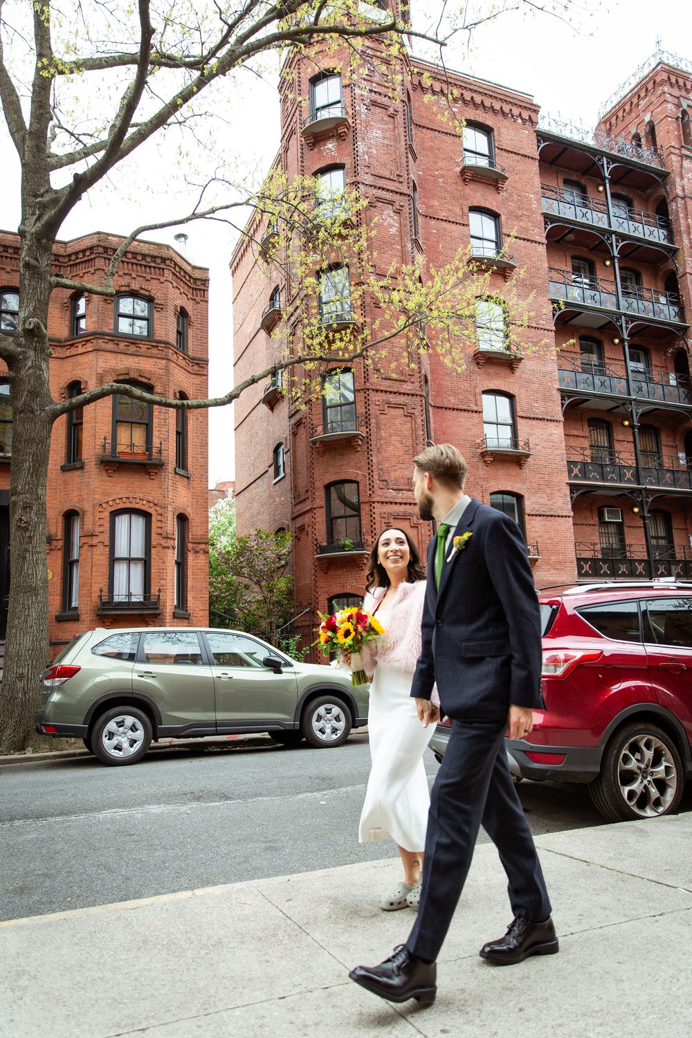 Bride and groom walking hand in hand on a city sidewalk with red brick buildings in the background.