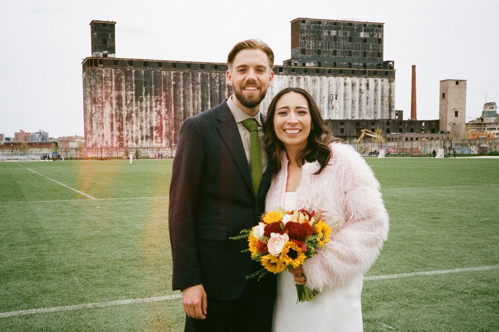 Couple poses on green field in front of industrial building, bride holds colorful bouquet in white dress and pink fur coat.
