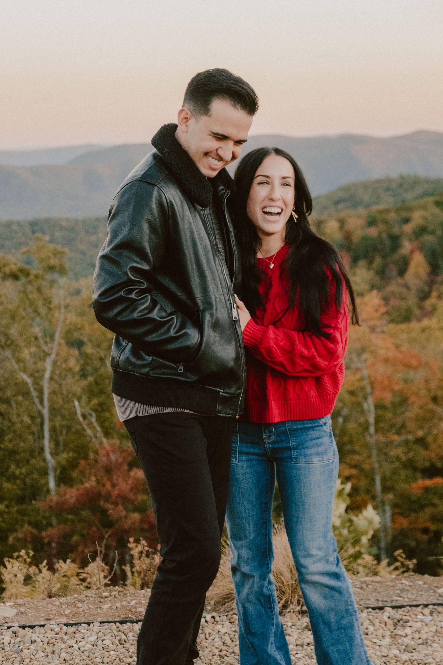 Couple laughing together on mountain overlook during autumn with fall foliage behind them.