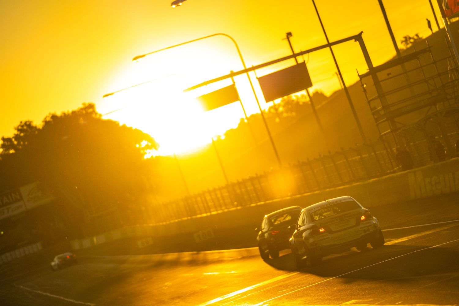 Race car drives along urban track during golden sunset with dramatic lens flare and silhouetted backdrop.