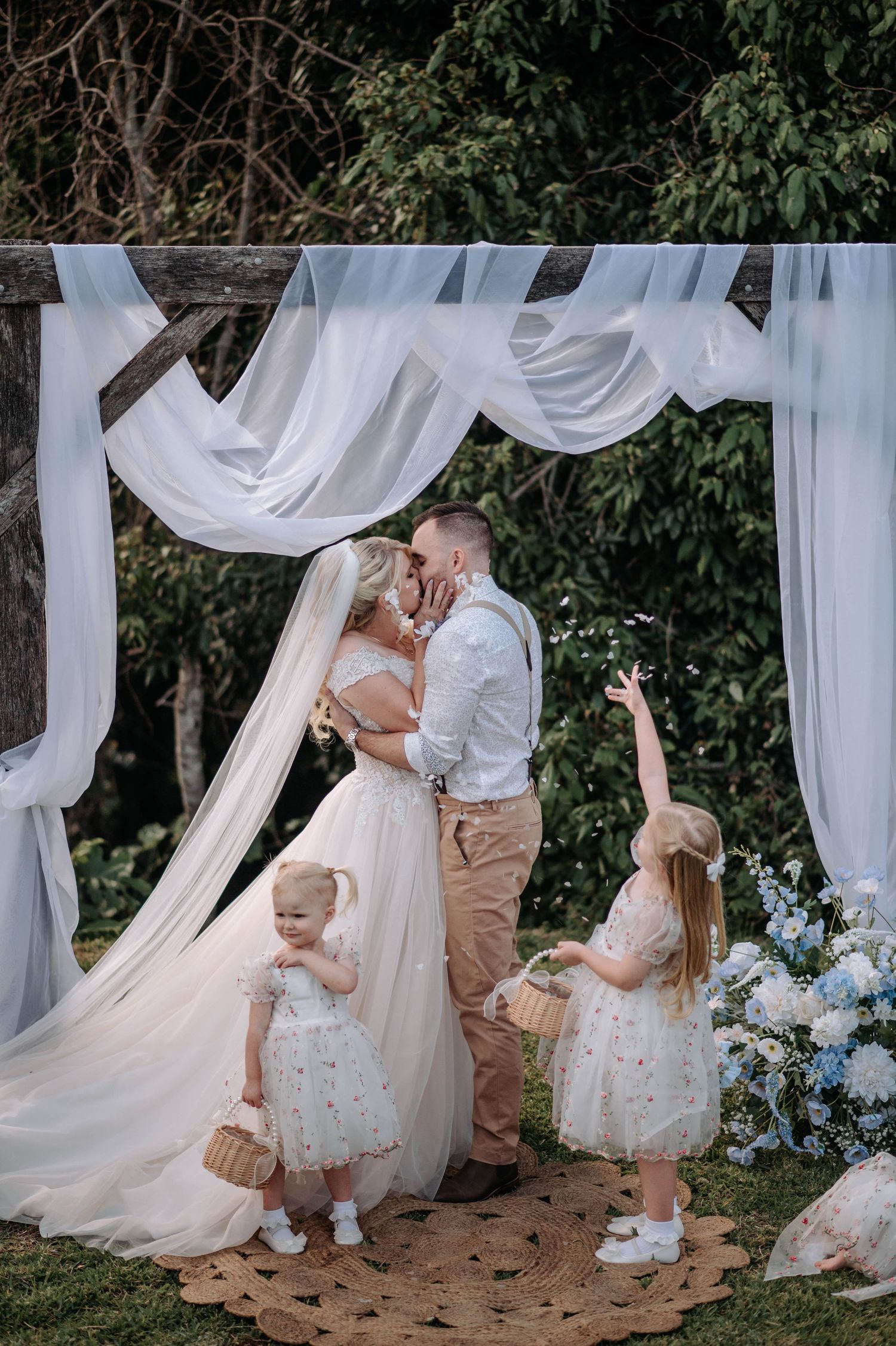 Wedding couple kissing under white draped arbor with children nearby.