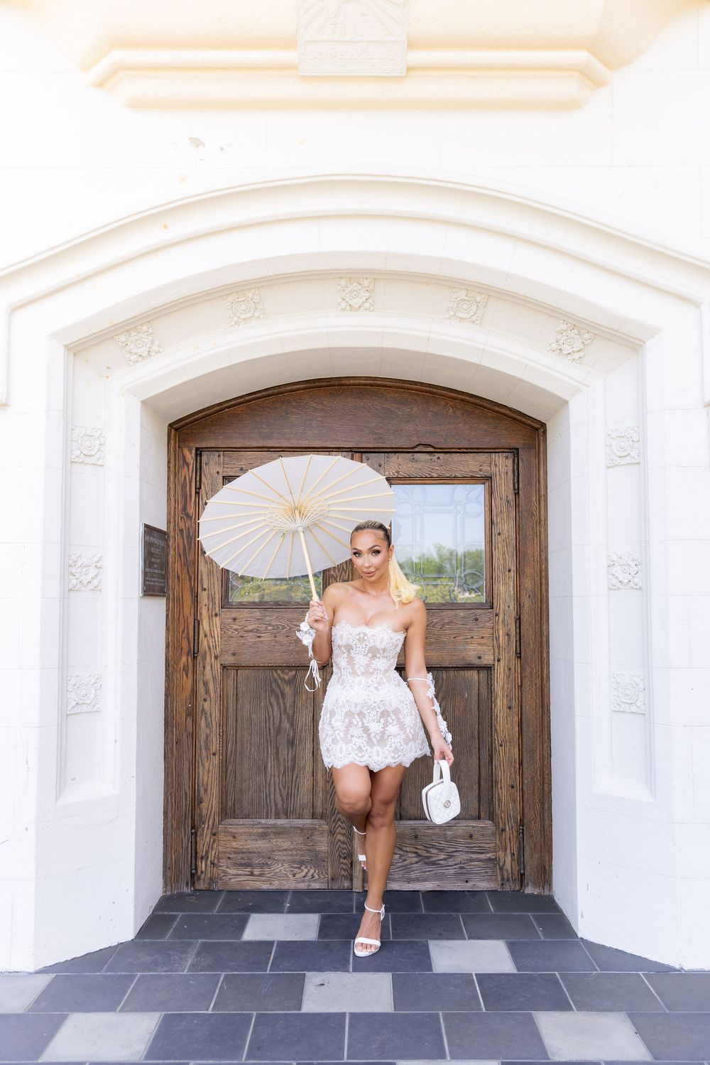 Elegant woman in white lace mini dress holds parasol while posing in arched wooden doorway.