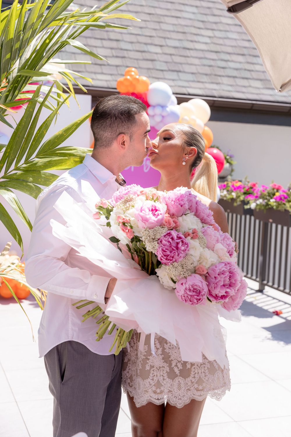 Couple shares a kiss outdoors holding large pink peony bouquet with colorful balloons in background.