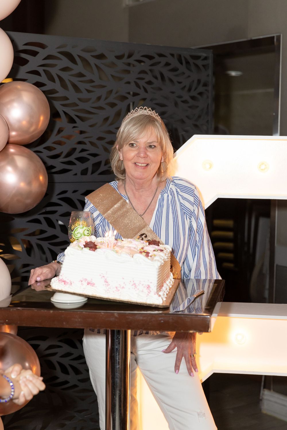 Smiling older woman wearing tiara and sash stands behind white birthday cake with balloons.