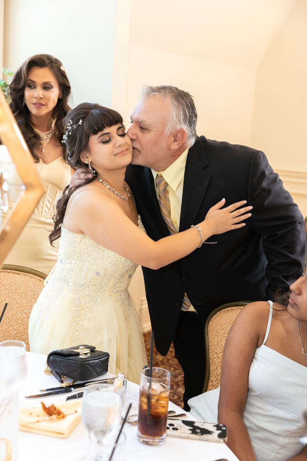 Older man in dark suit kisses bride on cheek at wedding reception dinner table.