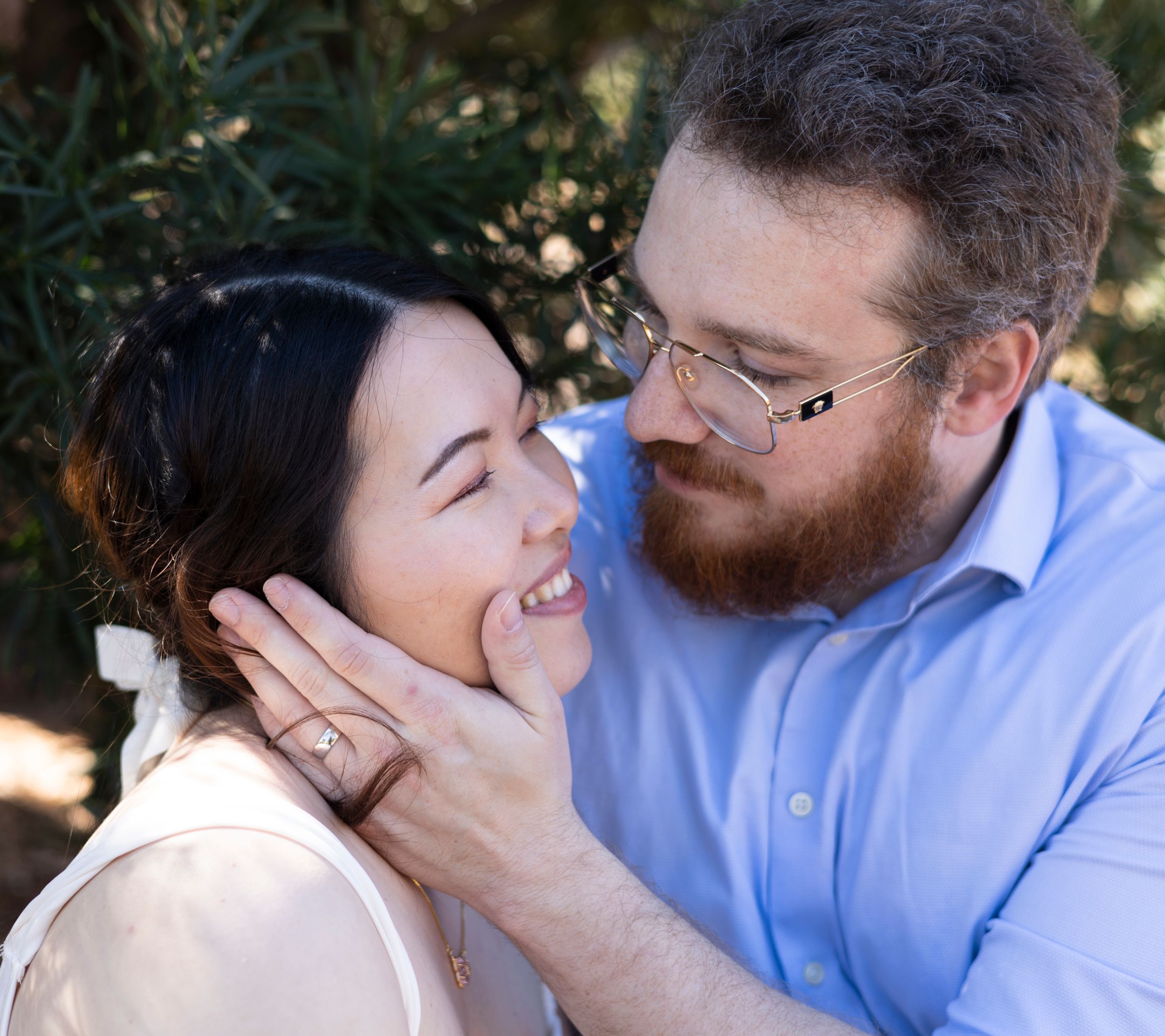 A happy couple shares a tender moment outdoors in natural lighting, with the person on the right wearing a blue button-down shirt.
