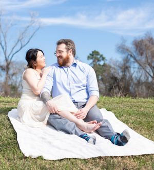 A couple sits together on a white blanket on a grassy hill during a sunny spring day.