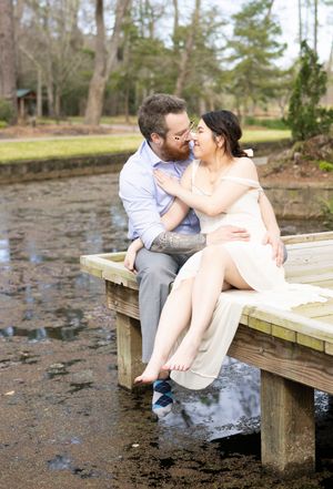 A romantic couple sits on a wooden dock by a lake sharing an intimate moment in a natural forest setting.
