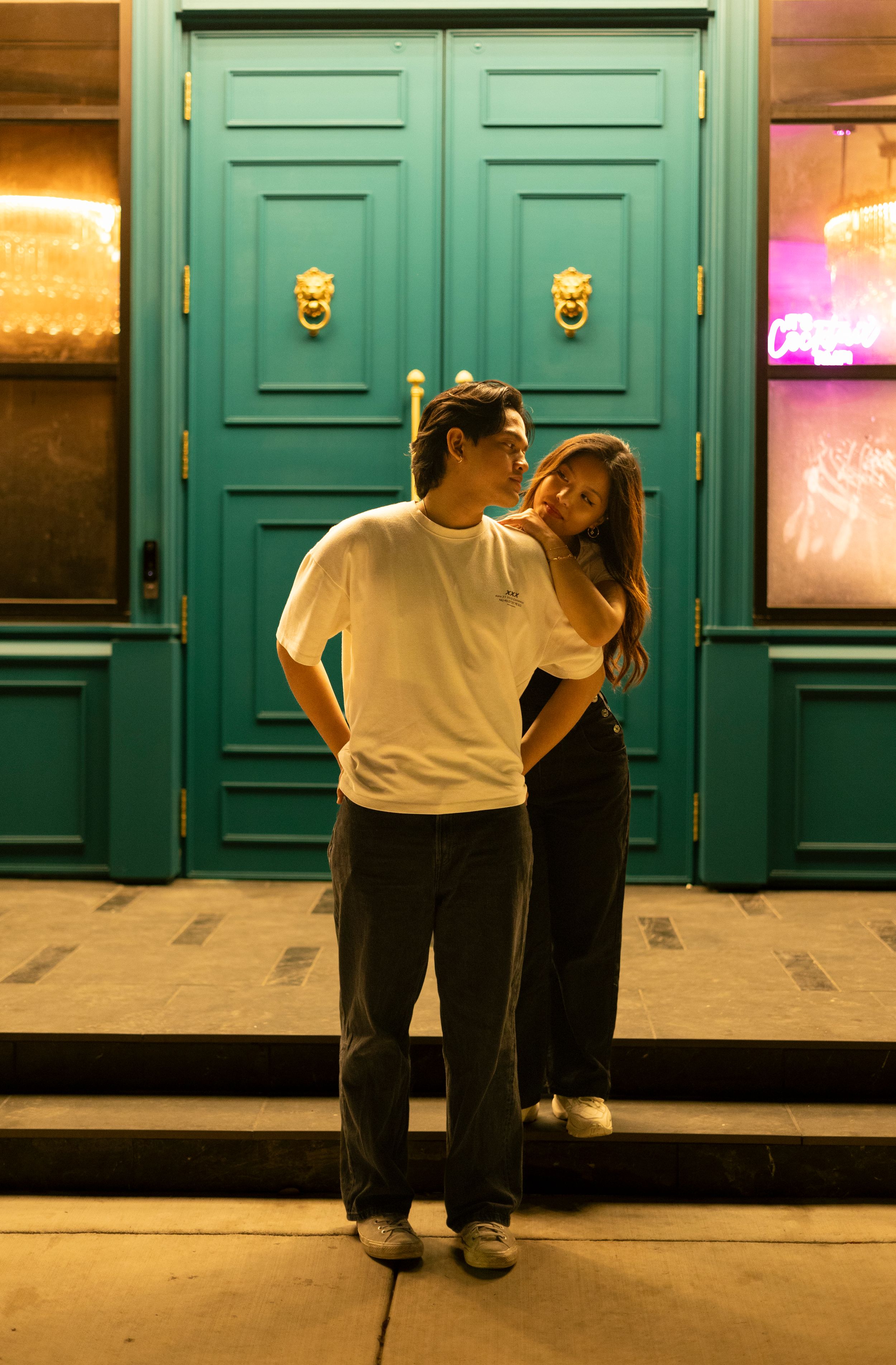 A couple embraces in front of turquoise double doors with brass lion knockers at night.