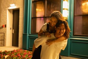 Two people share a joyful piggyback moment outside at night with warm lighting and flowers nearby.