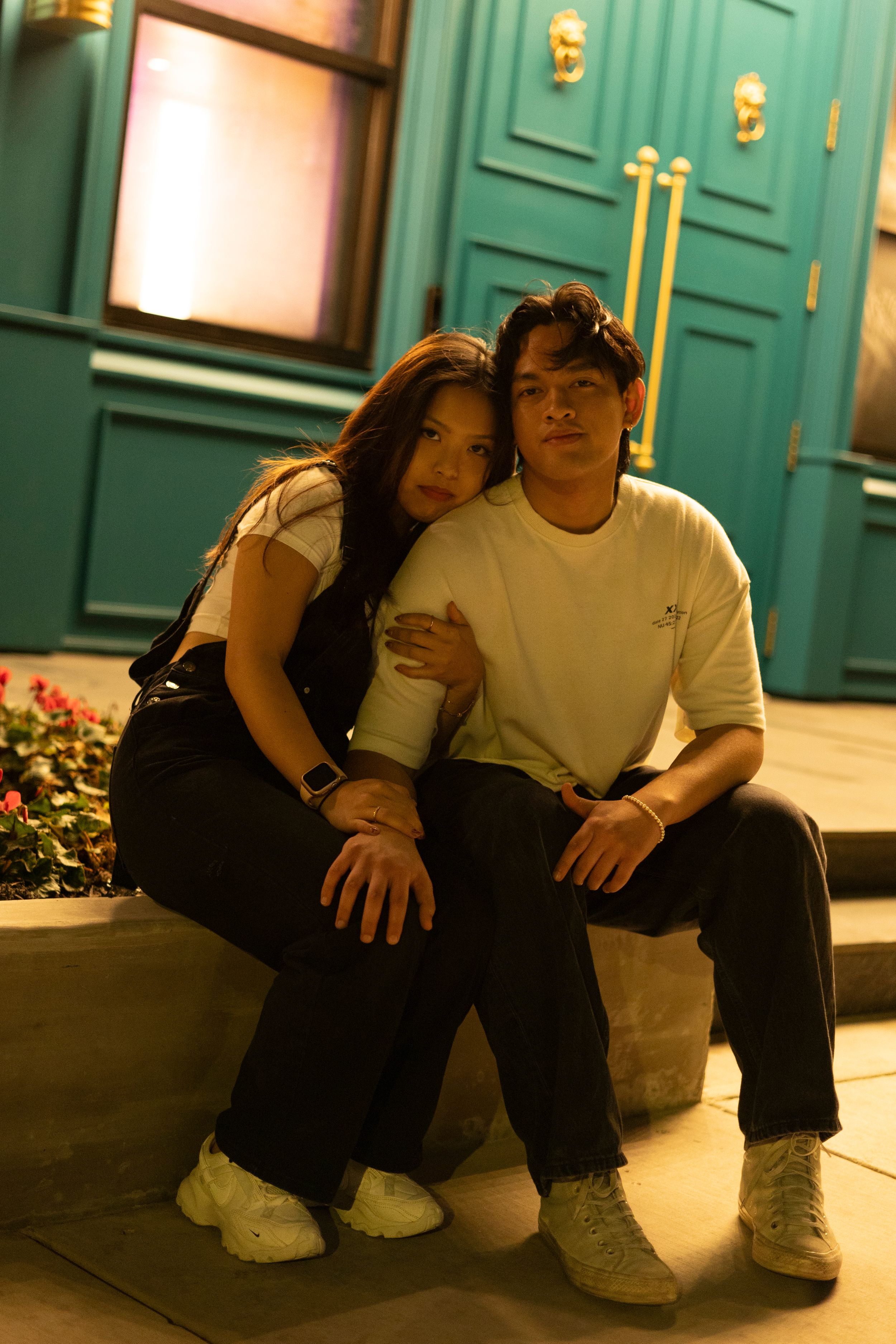 A couple sits together on outdoor steps at night in front of a teal-colored door with gold fixtures.