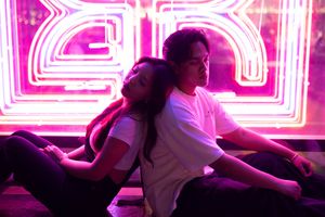 Two people relax together against a backdrop of vibrant pink neon lights creating geometric patterns.