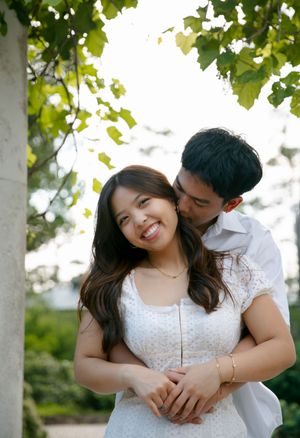 A couple shares a tender moment outdoors under green leaves in a garden setting, with the woman wearing a white lace dress.