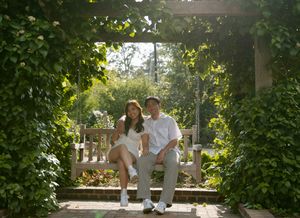 Two people in white outfits sit together on a garden bench surrounded by lush greenery and natural light.