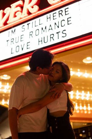 A romantic couple embraces beneath an illuminated theater marquee sign reading 'I'm still here true romance love hurts'.