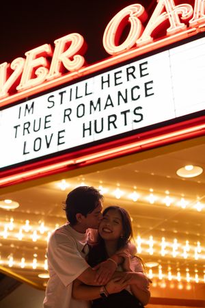 A romantic couple embraces beneath an illuminated theater marquee displaying 'I'm still here true romance love hurts'.
