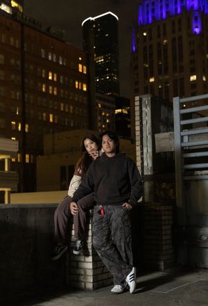 A couple stands together at night in an urban setting with illuminated skyscrapers and a blue-lit building in the background.