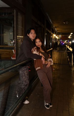 A couple poses together at night in a dimly lit urban setting with railing and glowing lights in the background.