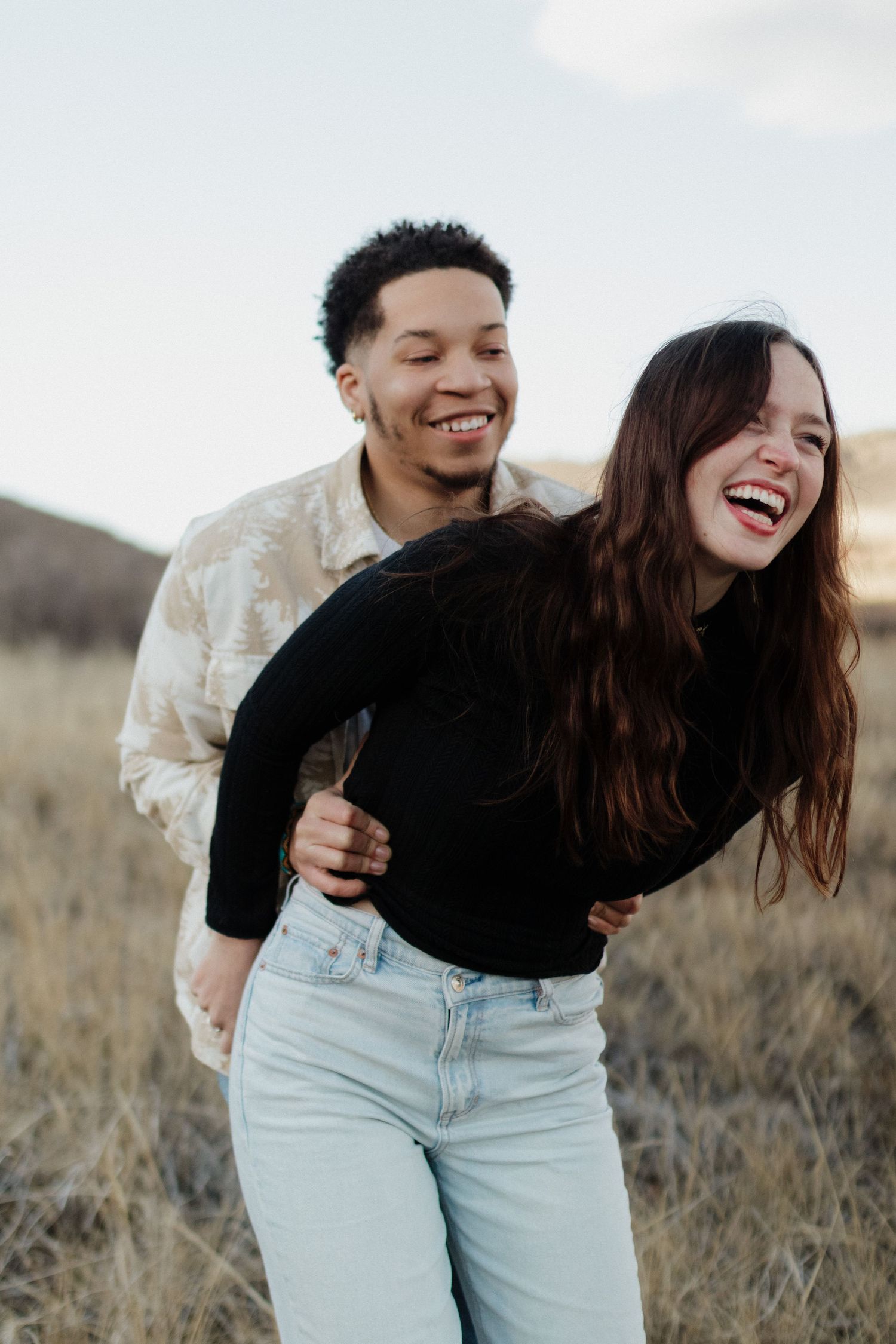 A couple laughs together while embracing in a field during golden hour with mountains in background.