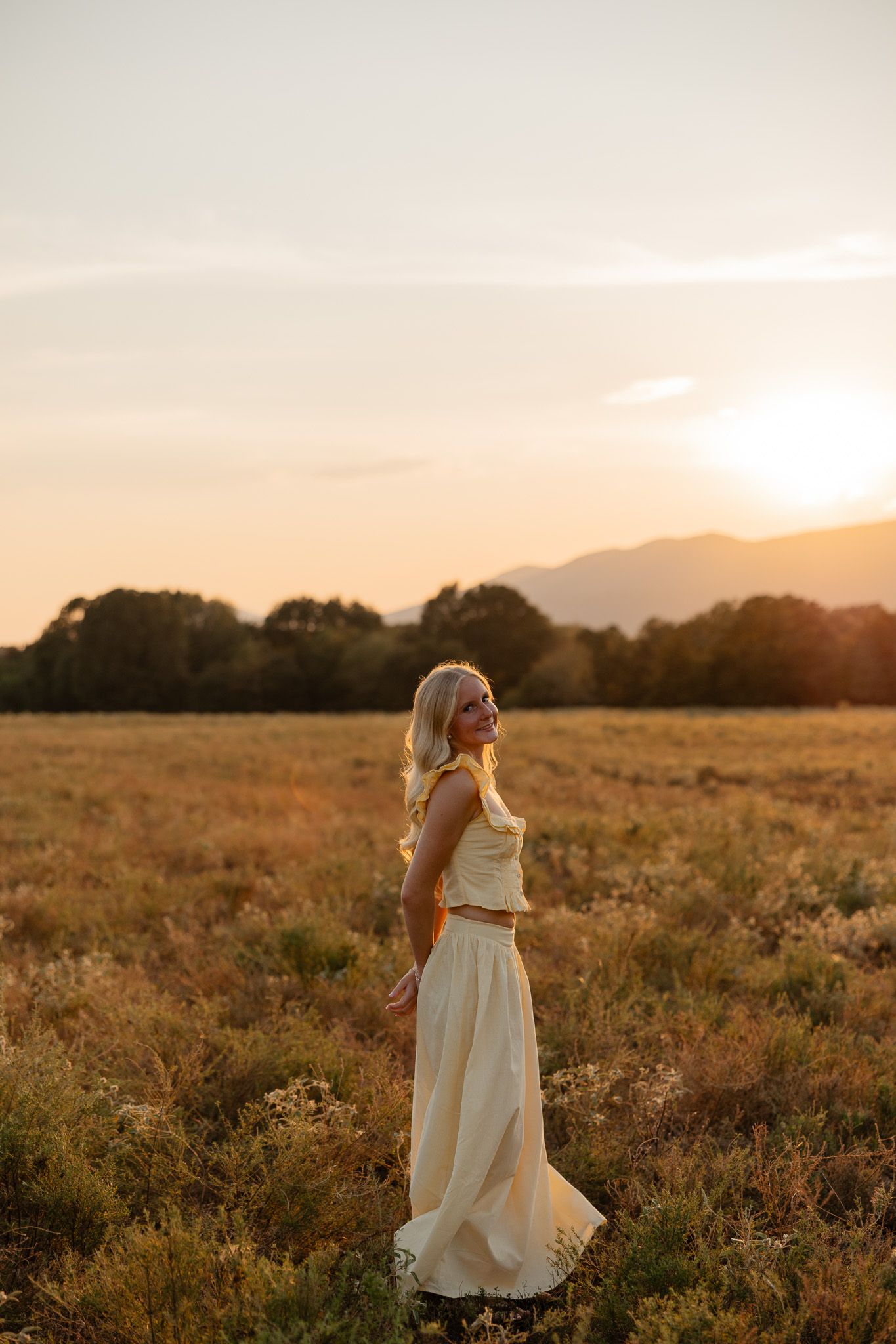 Woman in white dress twirls in golden field at sunset with mountains in background.