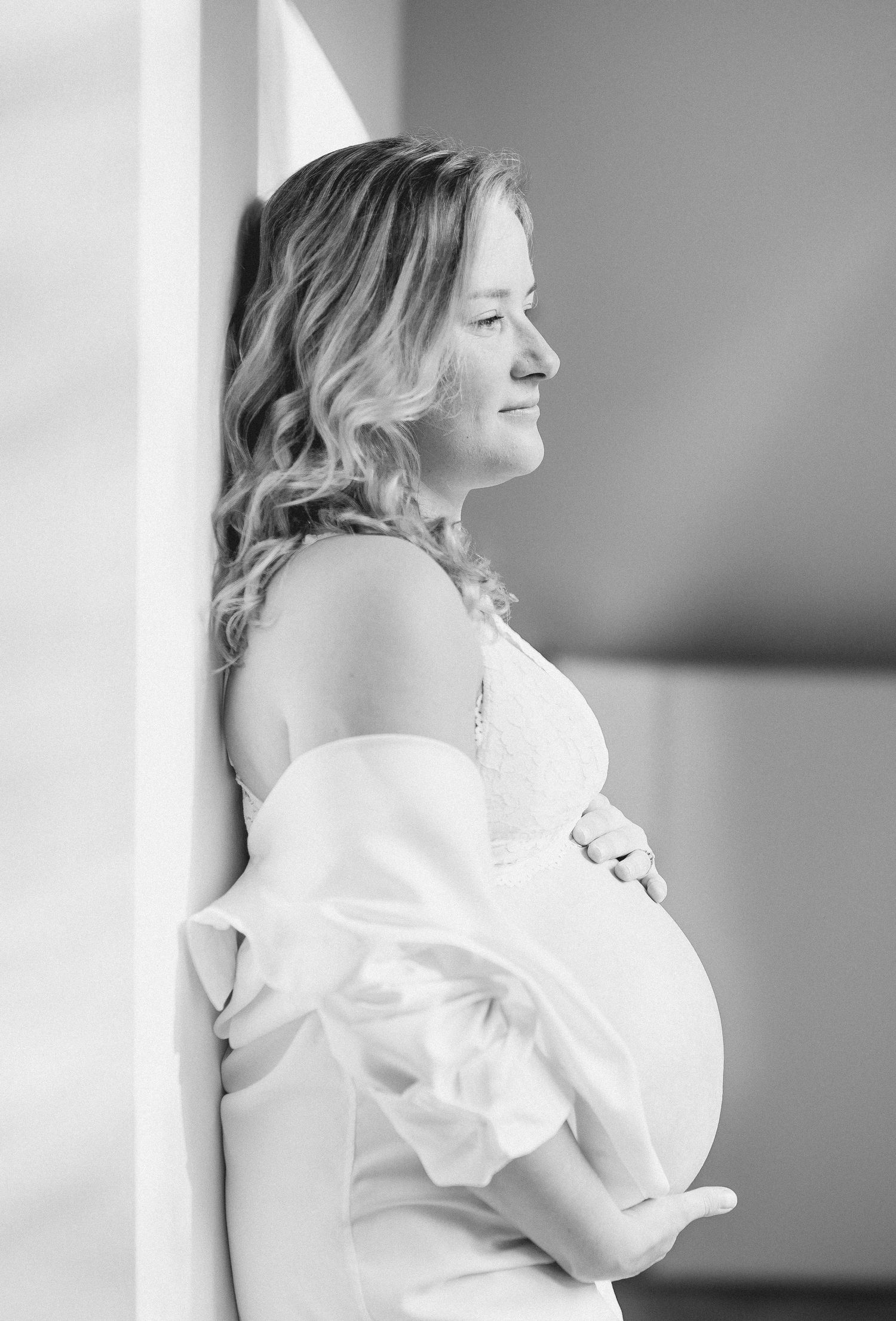 Black and white maternity portrait of an expectant mother in a white dress leaning against a wall - indoors photoshoot in Hoofddorp