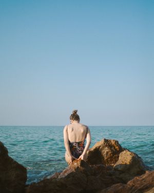 Person standing on a rocky cliff, gazing at the vast ocean under a clear sky.