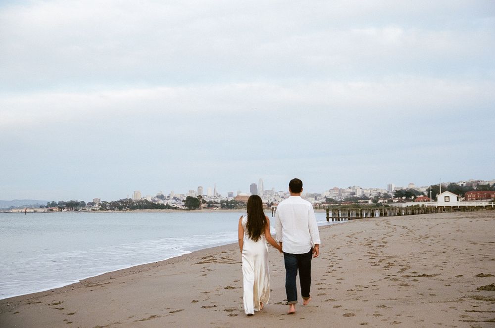 A couple walks together on a sandy beach with a seaside town and pier visible in the coastal background.