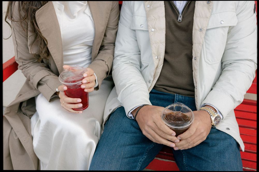 A couple sits on a red bench holding cold beverages while wearing casual clothing.