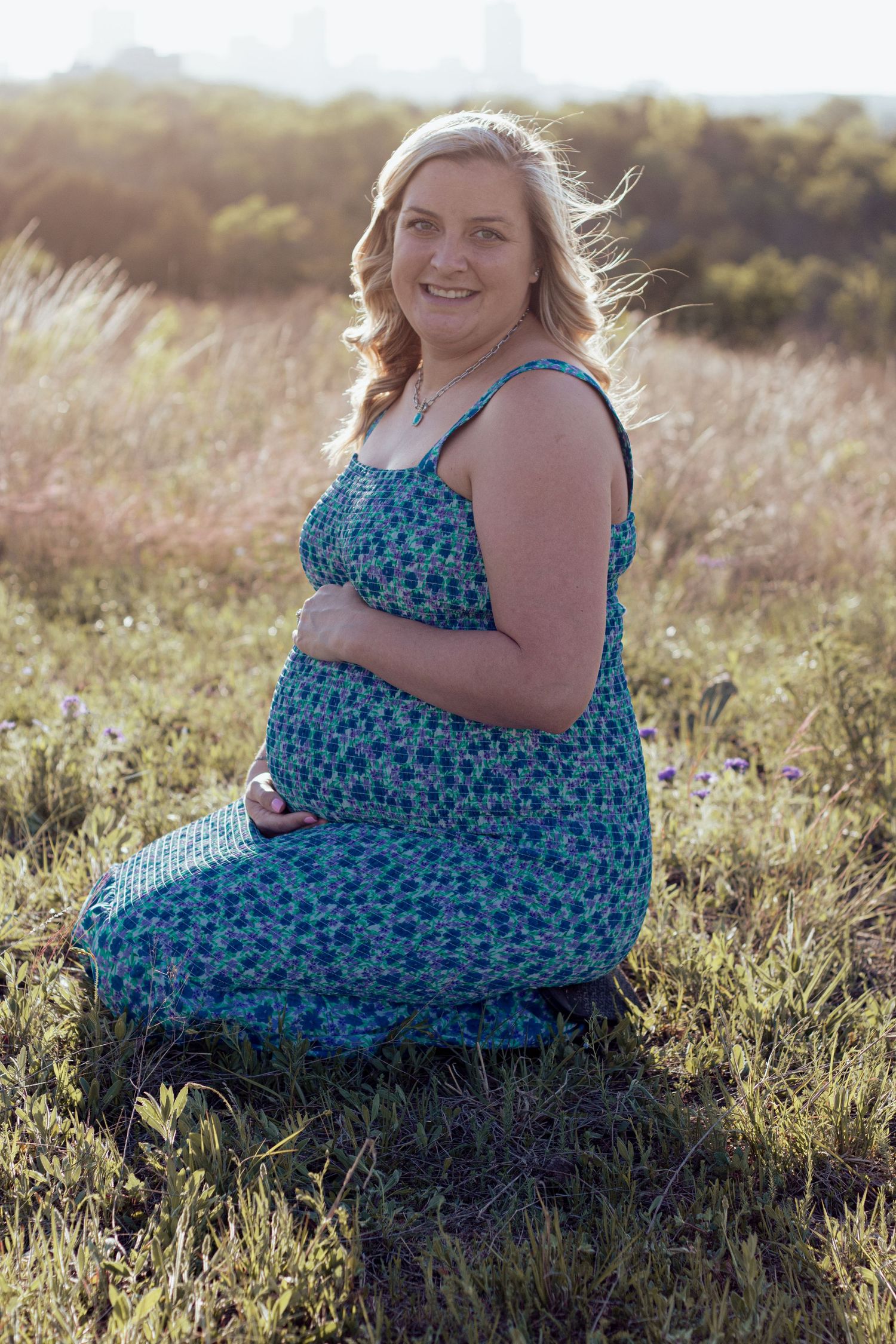 A person in a blue floral dress sits in a sunlit field of tall grass during golden hour.