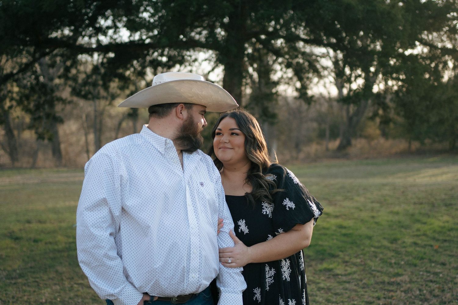 A romantic couple stands close together in a field at dusk, with the person on the left wearing a cowboy hat and white shirt.
