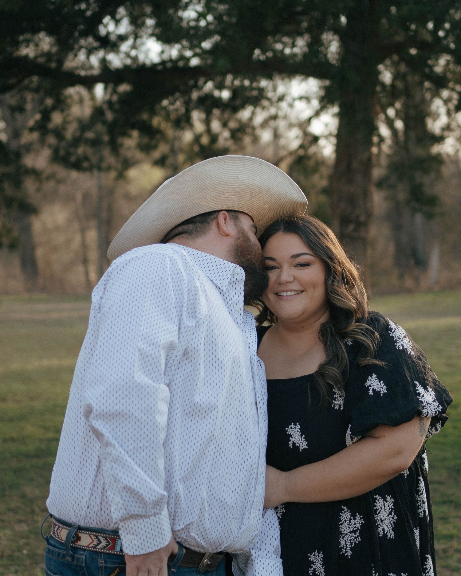 A romantic couple embraces in a natural outdoor setting with trees in the background at sunset.