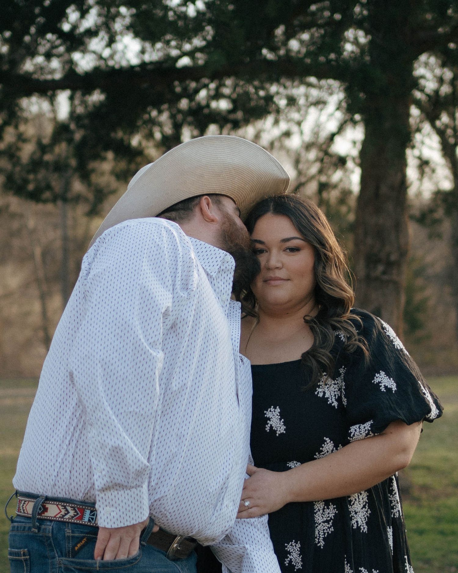 A romantic couple poses together outdoors at sunset in western attire with cowboy hat and floral dress.
