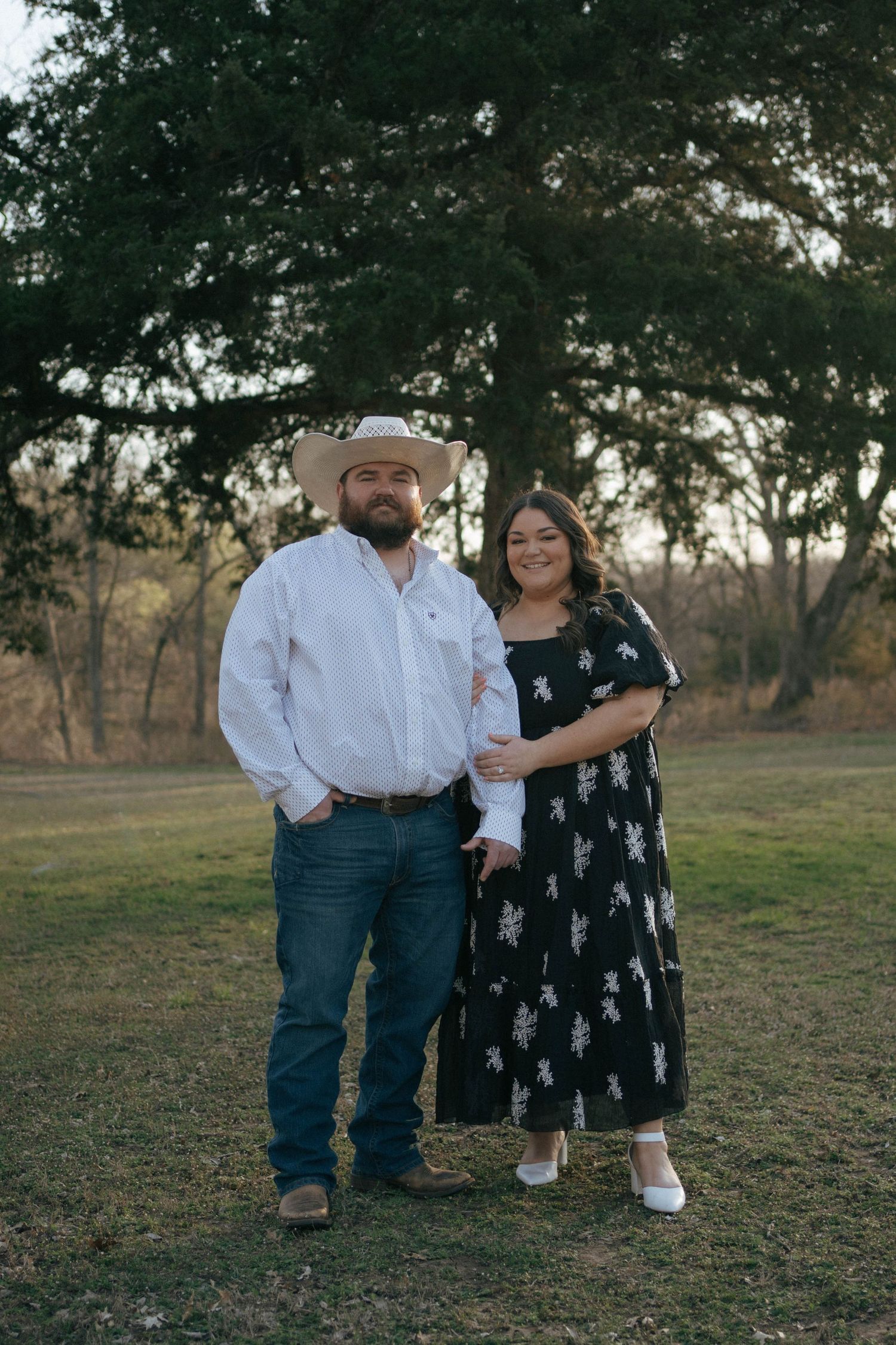A couple poses for a portrait outdoors in a field with trees creating a natural backdrop.