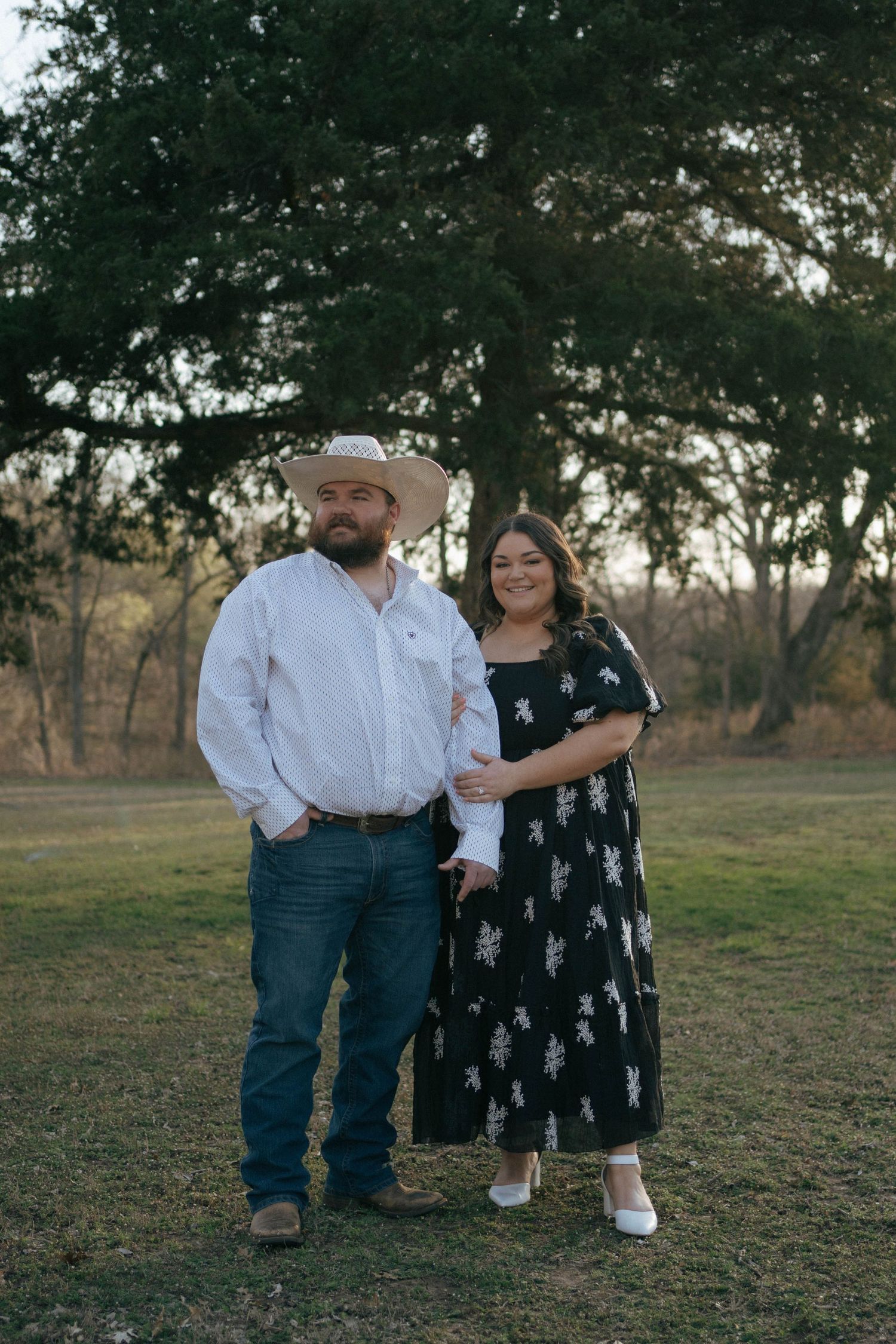 A couple poses together outdoors with trees in the background at sunset, wearing western attire.