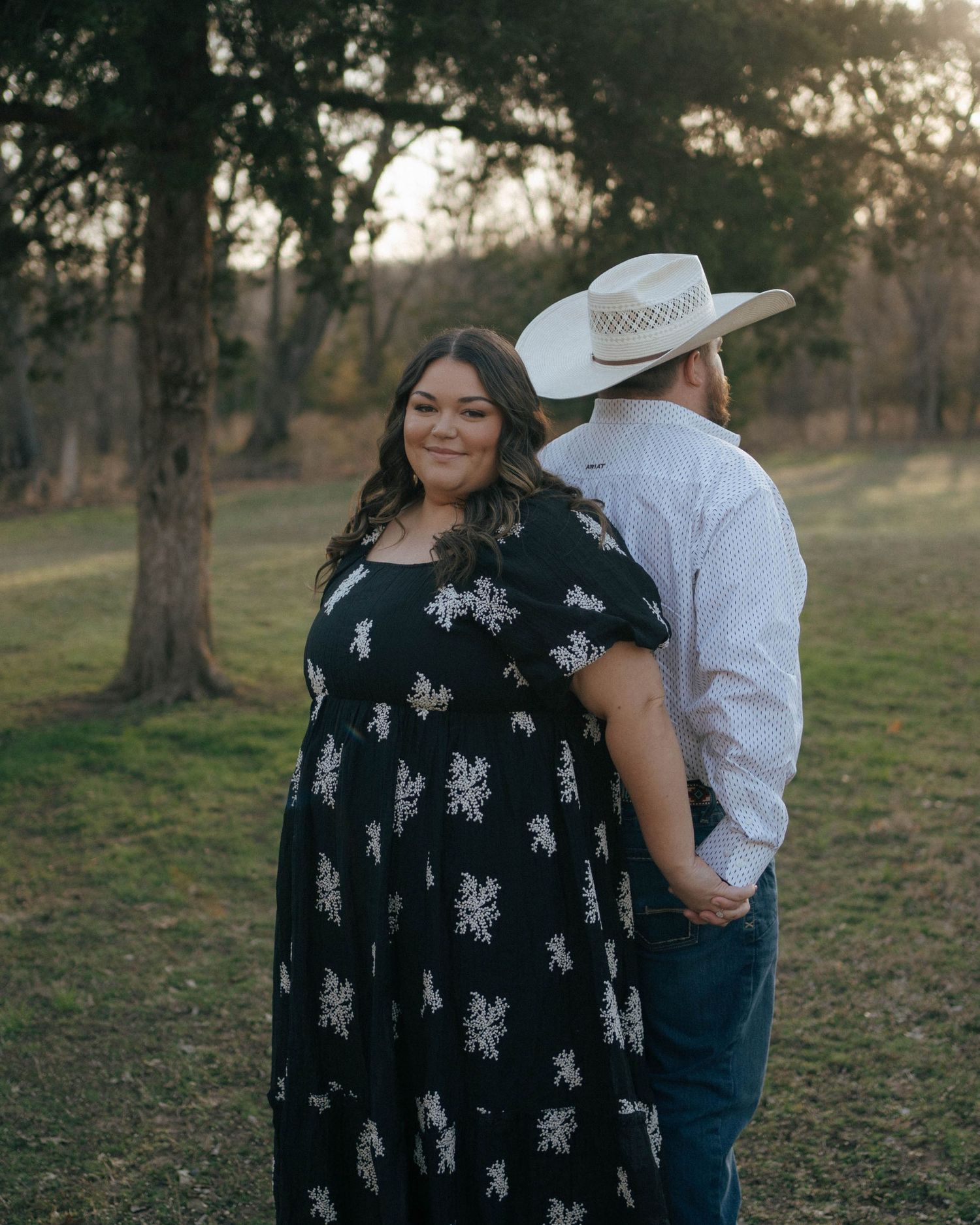 Couple posing in outdoor setting with trees at dusk, person in black floral dress and companion in white hat and jeans.