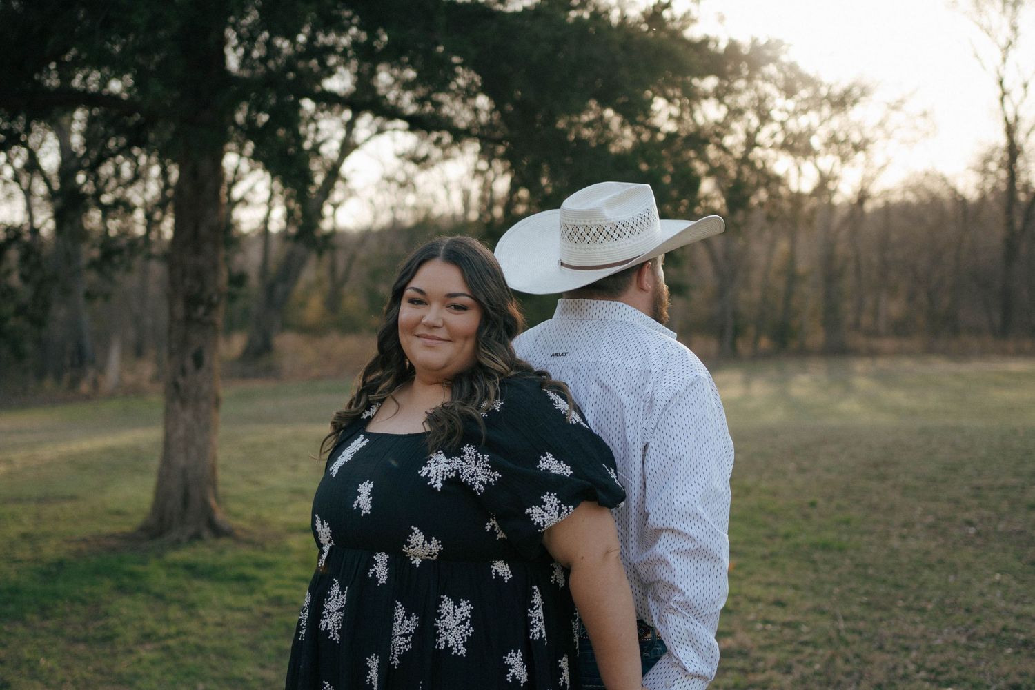 A couple stands together in a park setting during golden hour with trees in the background.