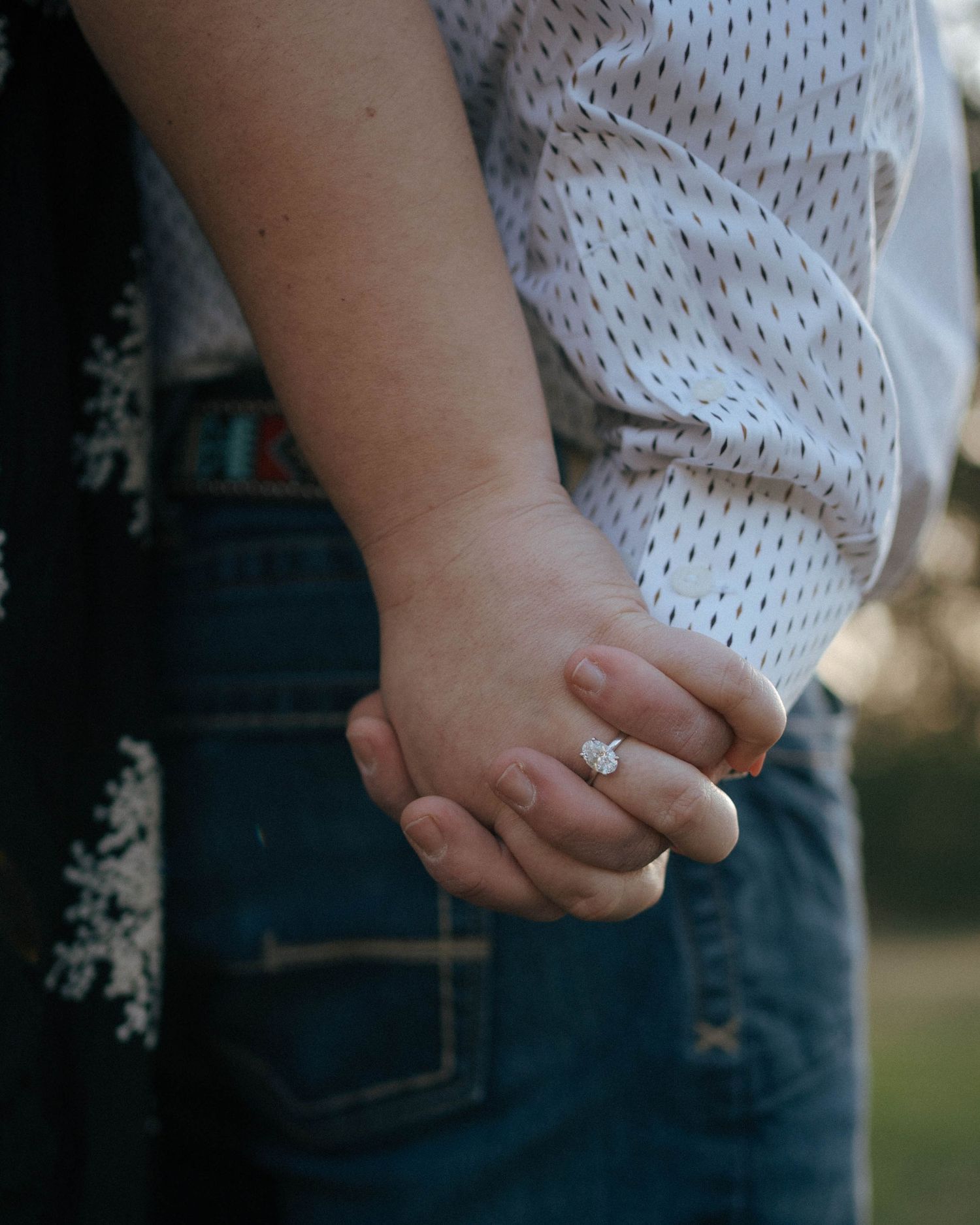 Close-up view of two hands clasped together with an engagement ring, wearing blue jeans and a white patterned top.