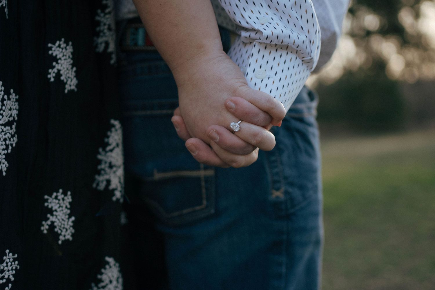 Close up shot of hands holding each other at sunset, wearing a ring and floral dress.