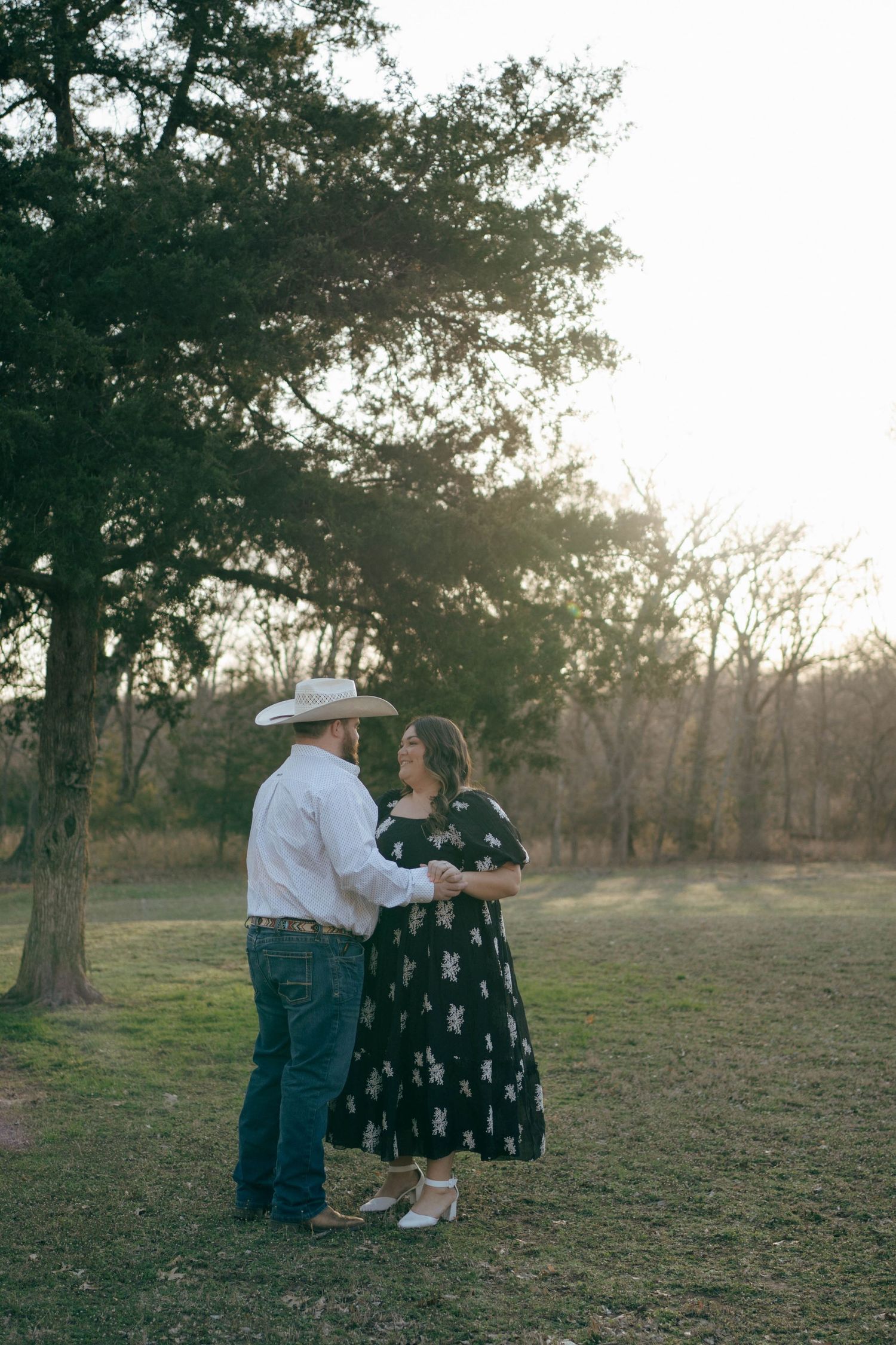 A couple in western attire dance together at sunset in a rustic outdoor setting among trees.